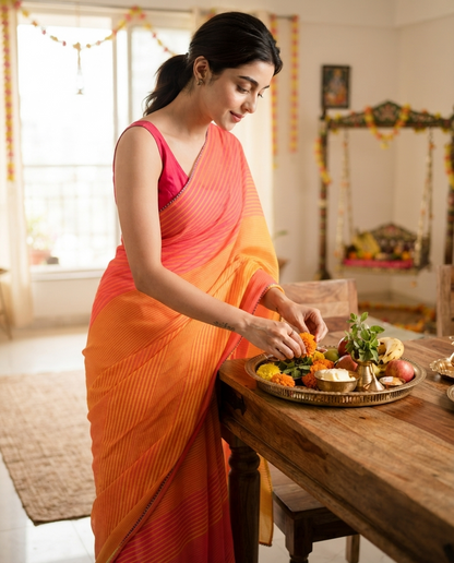 A woman in a vibrant orange and pink saree stands at a wooden table, preparing food from a brass tray during a traditional Indian festival. The background shows a warm, decorated home interior with a window and a hanging swing. karvachauth, karwa chauth