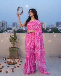 A woman stands on a rooftop terrace at dusk wearing a bright pink sari with white tie-dye stripes. She holds a small silver mirror in one hand and has a diya arrangement and potted plant beside her, with a city skyline in the background. karwachauth, karvachauth