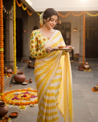 A woman stands in a decorated courtyard holding a tray with food, wearing a yellow and white striped saree with a floral printed blouse. rakshabandhan, festival