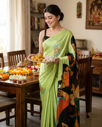 A woman wearing a bright lime green saree with colorful graphic prints stands in a decorated home interior, holding a plate of sweets. The background features a wooden dining table with flowers and traditional decor.rakshabandhan